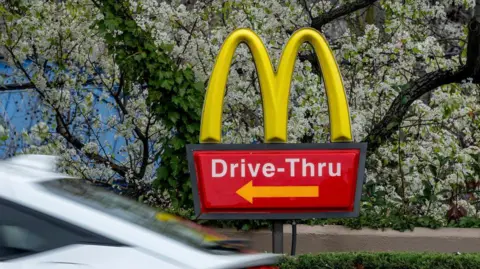 We can see a McDonald's 'Drive-Thru' sign. In the foreground, a car is passing by. Behind the red sign, with a gold letter "M", a tree is in blossom.