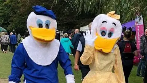 Maidenhead Lions Two people, one wearing a blue Donald Duck costume and the other wearing a yellow Daisy Duck costume. Daisy Duck is waving at the camera, and there's grass and crowds of people behind them.