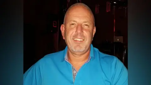 Tanned man in a blue shirt smiling at the camera. He has close cropped graying hair. He is in front of a dark background.