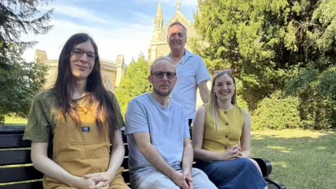 Amy Holmes/BBC Four people posing on and behind a wooden bench in a garden, with trees and a historic stone building in the background.