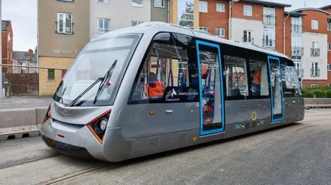 A very light railway carriage in Coventry, being run as part of a trial of a scheme. There are a number of people on board wearing orange high visibility jackets.