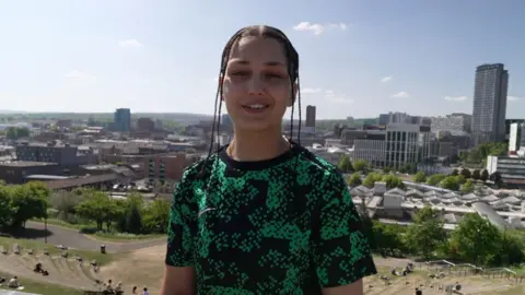 AJ is stood proud in front of the Sheffield landscape. She is wearing a black and green pattered T-shirt and her hair is in braids.