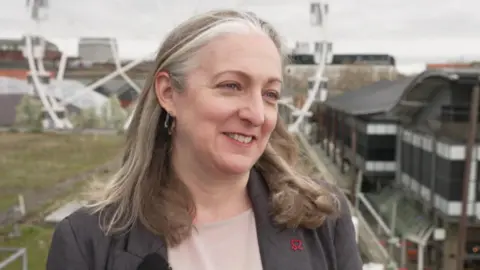 Danielle Poole, from the British Heart Foundation, she has shoulder-length brown and grey hair, and is wearing a grey top. She is smiling at the camera, and is interviewed on the roof of a central city structure