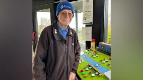A man in a blue hat and grey coat with a white beard, operating a mechanical lock 