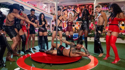Peacock via Getty Images A group of people in leather outfits stand around a pole in the backyard of a large house. This is part of a challenge the Love Island contestants participated in.