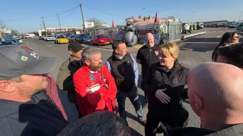 A woman in a dark jacket with a Unite union logo gestures as she talks to a group of men gathered around her in a semicircle. Behind them, an inflatable grey rat stands by metal depot gates alongside red flags and protest placards. The scene is set on a sunny day in an industrial area with parked cars and commercial vehicles visible in the background.