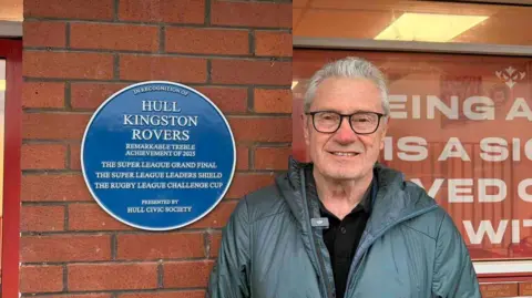 A man with white hair, he has a grey goat on and is smiling at the camera wearing glasses. He's stood outside of Craven Park's stadium next to the new blue plaque marking Hull KR's achievements. 