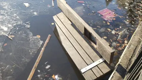 The remains of a broken wooden bench floating on the surface of a body of water surrounded by pieces of broken ice. 