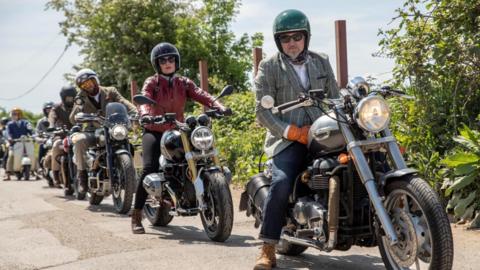 A line of men people dressed in suits sitting on their motorcycles on a country lane