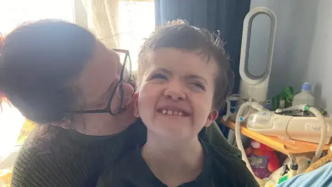 BBC Michelle Lloyd smiling and speaking into her son Billy's ear as she dresses him for the day