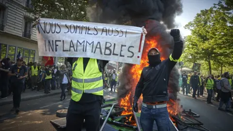 Getty Images Gilets jaunes (yellow-vests) are seen standing in front of a burning barricade with a banner reading 'we can also burn' on 20 April