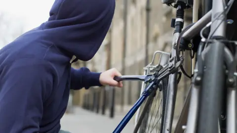 A person in a blue hoodie attempts to chop off a bicycle lock with bolt croppers.