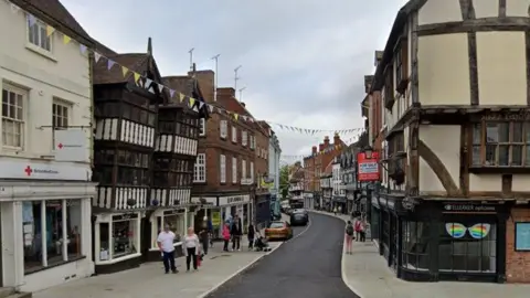 Generic view of a town centre street with a mixture of timber-framed and brick buildings. Several cars are parked in bays at the side of the street, and people are walking along the pavements.