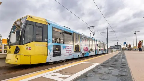A yellow and blue Metrolink tram at Trafford Palazzo on a cloudy day.