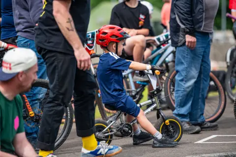 Pete Scullion A little boy in a blue top and T-shirt sits on his bike with a red helmet on
