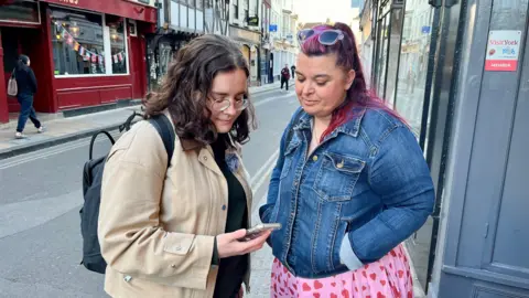 Two women stand on the pavement in a street in York, looking at a mobile phone.