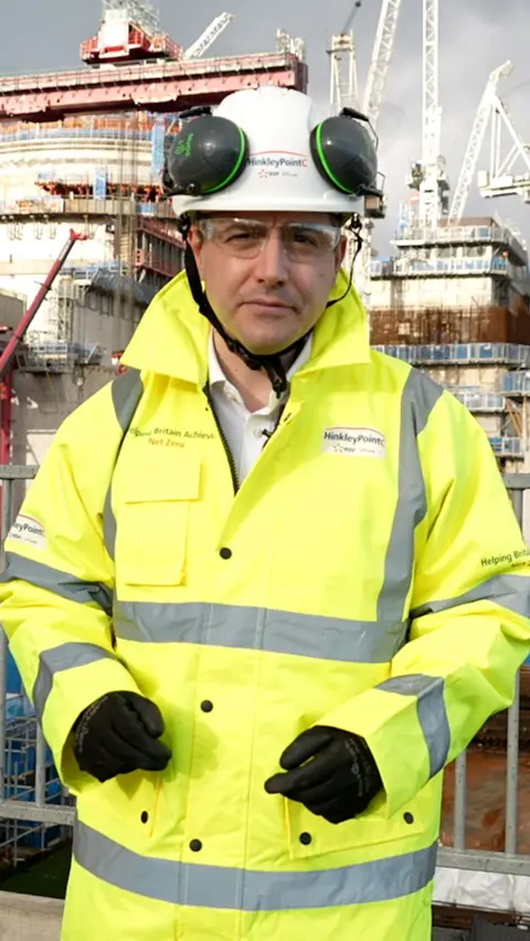 A man wears a yellow hi-vis jacket and white helmet. He is standing in front of a construction site and looking down the camera.