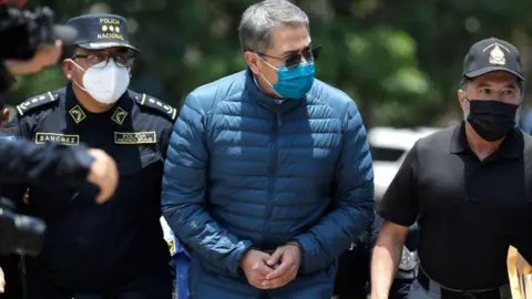 Photo by Jorge Cabrera/Getty Image Juan Orlando Hernández, wearing a blue puffer jacket, a Covid mask and dark sunglasses, is escorted by police officers ahead of his extradition to the US on 21 April, 2022 in Tegucigalpa, Honduras. His hands are shackled.