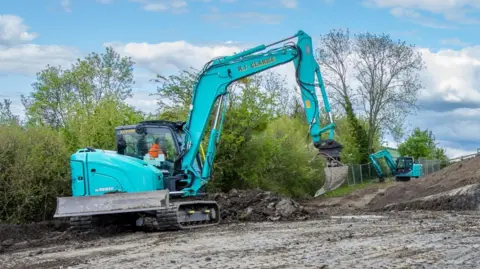 Ian Crowder A blue 'R J Clarke' excavator is digging into mud. It is being operated by a person wearing an orange high-vis jacket. 