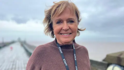 Nicole Laken. She has short brown hair and is wearing a brown jumper and a black lanyard with "staff" printed on it. She is pictured outside on Clevedon Pier. Her hair is blowing in the wind. She is looking directly at the camera and smiling. 