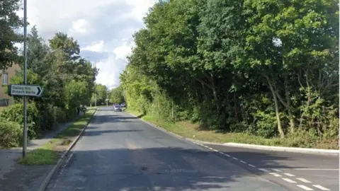 Google Road junction lined by trees. A sign on the left points right to Galley Hill and Breach Barns. There is an unlined main road and dotted lines across the mouth of the road to the right.