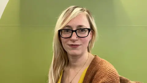 A woman with long blonde hair and glasses sitting on a chair.