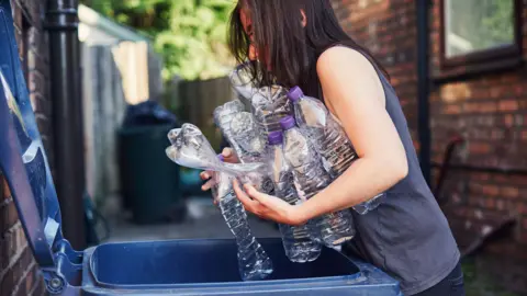 A woman in a sleeveless grey vest with her arms full of empty plastic bottles, leaning over an open blue bin. She is in an alleyway and her dark hair is falling over her face. It is a sunny day and brick buildings and wooden fences can be seen behind.