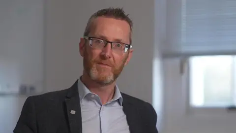 A man with short, dark hair spiked at the front. Wearing black glasses, a grey blazer and blue shirt. He is pictured against a cream wall with a window behind him.