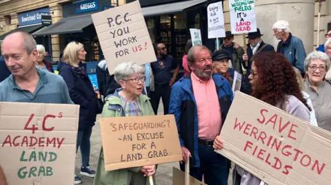 Shariqua Ahmed/BBC Several people holding home made placards stand on a pedestrianised street. Placards say "Save Werrington Fileds" "Safeguarding? An excuse for a land grab" and "PCC you work for us"