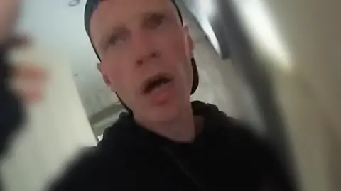 Police body-camera footage of Kevin Weetman, wearing a baseball cap with the peak backwards and a dark hoodie, looking at the officer in shock