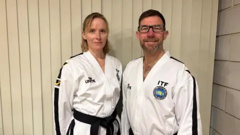 A man and a woman dressed in martial arts uniform, with black belts smile at the camera. On the left is a woman with shoulder length blond hair and on the right is a man with brown hair, a beard and glasses.