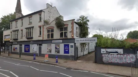 Google A three-storey building mainly on the left of the photo has the Dulux logo on the outside at the top of the ground floor. A road is at the bottom of the image and a sign with the words car park entrance is near the middle of the photo.