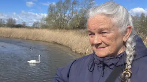 BBC/Lara King A woman with long grey hair in a pony tail and wearing a blue coat. She is standing in front of a swan which has black oil on it, a swan is visible swimming in a lake and there are golden reed bushes surrounding the water.