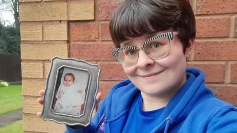 Elizabeth Rees-Gilbert has short dark brown hair. She is wearing striking glasses with a black and white striped frame. She is outside wearing a blue hoodie and holding a framed picture of herself as a child