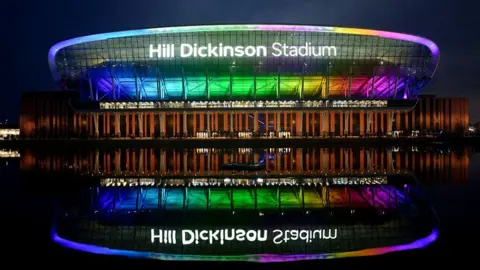 External view of Everton FC's Hill Dickinson Stadium at Bramley-Moore Dock lit up in rainbow colours ahead of an evening Premier League game.