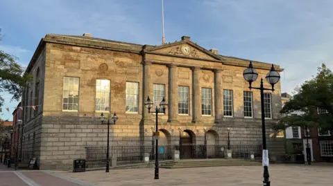 Shire Hall in Stafford, a stone building with a clock at its centre above four central pillars. There is an open public square and a number of ornate lampposts in front of the building. There are nine large windows on the building and black railings immediately in front of it.