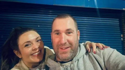 Ritchie Massey pictured with a woman who is smiling and has her brown hair in a ponytail. Mr Massey has short dark hair, grey stubble and wears a grey hoodie while stood in front of blue shutters.