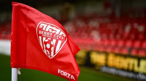 A red corner flag with the Derry City FC emblem is pictured inside the Brandywell