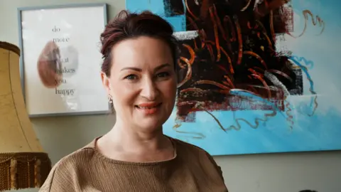 John Fairhall/BBC A head and shoulders image of a woman wearing a brown top while standing in front of an artwork on the wall. She is looking into the camera.