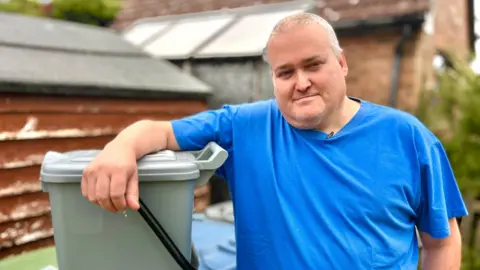 Martin Giles/BBC Carl Creed is in a blue T-shirt and is standing in a garden with a brown shed in the distance. Carl is leaning his arm over his grey food caddy waste bin a which has a black handle. 