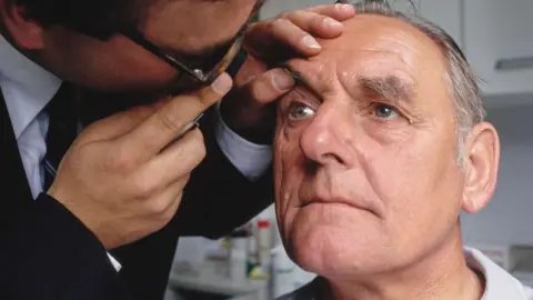 A man with grey hair having his eyes tested by another man who looks into his eyes with an ophthalmoscope 