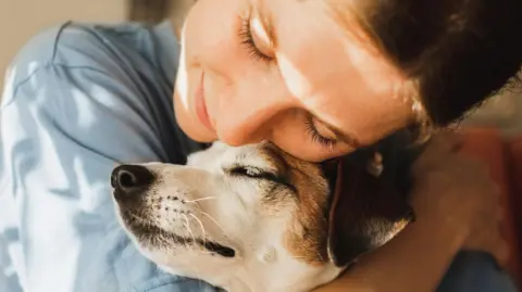A young woman wearing a blue stop cuddling her Jack Russell Terrier. 