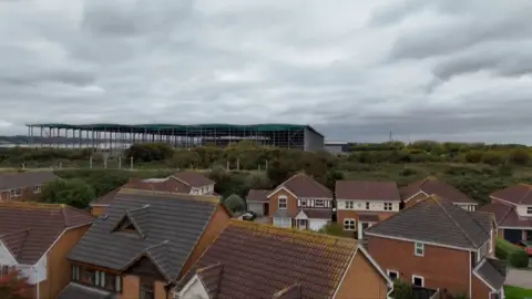 The image shows a giant warehouse being built on land next to the Severn Estuary but close to houses in Pilning.