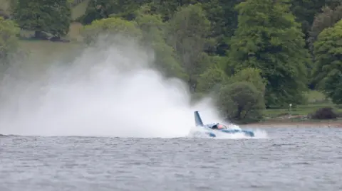 Matthew Willis Bluebird - a craft that looks like a cross between a plane and a boat - leaves a plume of spray behind as it moves across Coniston Water. Trees fill the background.