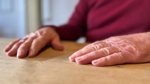 The older mans hands on a wooden table, blurred red jumper behind