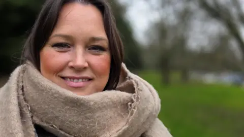 Paul Moseley/BBC Lucy Shires is standing on grass in front of some trees. She's smiling and wearing a large beige scarf.