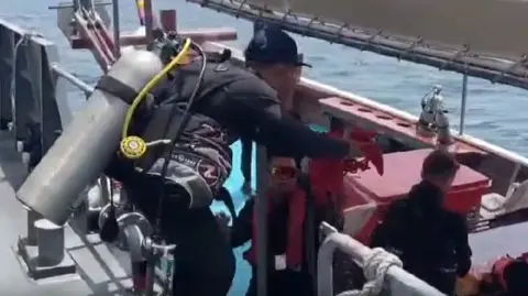 Royal Thai Navy A man on a ship, wearing black and carrying an air tank and equipment on his back, bends down to hand red equipment to another man on a smaller ship docked next to it