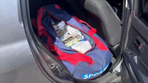 Lincolnshire Police A branded Sports Direct duffel bag with illegal vapes and cannabis cigarettes inside. It is on the back seat of a black car - the car door is open. 