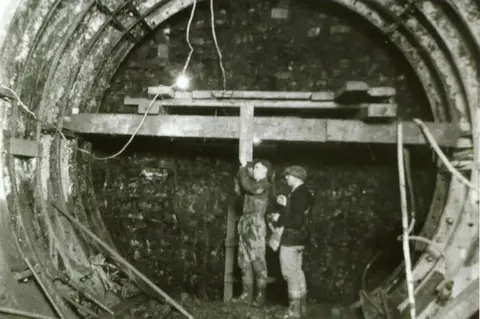 Brett Exton A black and white photo of two men building a tunnel, a brick wall is visible at the end and a metal tube is. There is tubes and wires hanging down. 