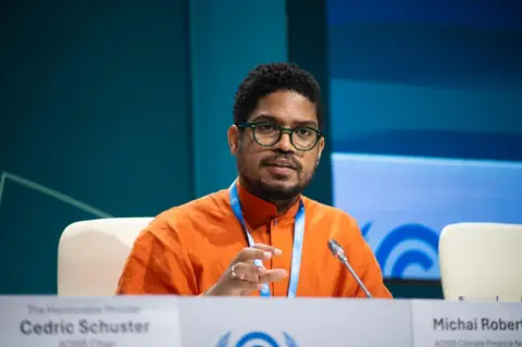 AOSIS Michai Robertson speaking on a podium, wearing an orange shirt